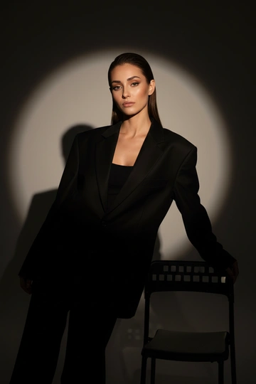 Модная фотосессия в студии, женщина одета в чёрный свободный пиджак, волосы гладко зачёсаны назад, профессиональный макияж, один прожектор, тёмный фон с круглым световым пятном, чёткая тень за спиной, кинематографичный свет, драматичный минимализм, модная съёмка для журнала, очень реалистично, средний и крупный план, лицо в фокусе, детализированная текстура кожи. женщина слегка отклонилась назад, одной рукой опирается на стул, свет подчёркивает ключицы и линию плеч, средний план