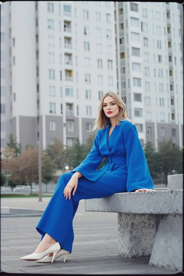 Женщина в ярко-синем трикотажном костюме и белых туфлях, широкие рукава, пояс на талии, позирует сидя на бетонной скамейке возле типовой советской высотки, взгляд в камеру, атмосфера полароидной фотографии конца 70-х, мягкий уличный свет.