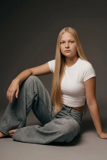 Студийная фотография этой женщины. Не меняй внешность, черты лица должны оставаться такими же, как на исходном фото. На ней белая укороченная облегающая футболка в стиле 90-х, очень широкие свободные baggy серо-голубые джинсы как на референсе. Образ повседневный, чистый, современный. Волосы ухоженные, прямые, блестящие, очень длинные, ниже пояса..  Фон — однотонный тёмно-серый студийный бесшовный градиент без текстуры, с мягким переходом от более тёмного верхнего фона к чуть светлому у пола. Освещение — мягкий студийный свет: основной источник света расположен спереди и чуть сверху, свет рассеянный, без жёстких теней, лёгкий объёмный свет подчёркивает форму тела и складки одежды, тени мягкие, естественные, падают под ноги, кожа выглядит натурально, без пересветов и глянца. Контраст умеренный, цветокоррекция теплая, с слегка приглушёнными цветами, похожими на сепию.  Общее ощущение света — спокойный, редакционный, high-fashion studio look. Профессиональная съёмка, чистая композиция, реалистичное качество, без искажений пропорций и лица. Женщина сидит на полу боком к камере. Одна нога согнута и подтянута к корпусу, вторая согнута и уходит в сторону. Одна рука опирается о пол за спиной, вторая расслабленно лежит на бедре или колене. Корпус слегка наклонён назад. Голова повернута в сторону, взгляд направлен в камеру, выражение лица сосредоточенное и спокойное.