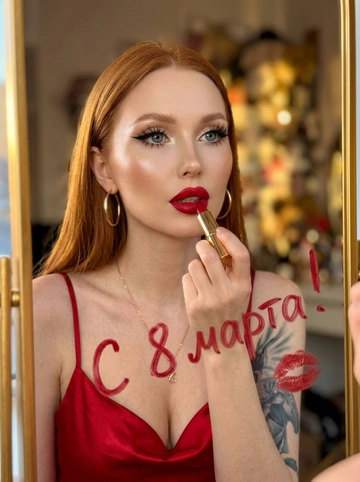 Отражение женщины в зеркале: женщина держит красную помаду. Гламурный макияж глаз с длинными чёрными стрелками, длинные густые ресницы с эффектом «лучики». Хайлайтер во внутренних уголках глаз и на скулах. Губы накрашены этой же красной помадой. Красное атласное платье на тонких лямках. Текст написанный на зеркале помадой от руки на русском языке без ошибок. Знак поцелуя красной помады на зеркале. Передний план: женское лицо, верхняя часть тела, рука, держащая помаду. Фон: зеркальная поверхность с текстом и поцелуем, размытое отражение интерьера. Глубина: неглубокая глубина резкости, фокус на женщине и поверхности зеркала. Портрет крупным планом, отражение, глаз, портретная линза, неглубокая глубина резкости. Тёплый золотой свет «золотой час», сильное боковое освещение. Женщина наносит помаду, отражение в зеркале, отражение в помещении, в помещении. Кожа, атласная ткань, золото (серьги, корпус помады, зеркальная рамка), стекло (зеркало). Фото сделано на iPhone 17 Pro Max.


Какая будет надпись?: Текст написанный на зеркале "С 8 марта!"