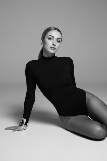 Стильная фотосессия в светло-серой студии с мягким рассеянным светом. Женщина одета в черный боди с длинными рукавами и высоким воротником. На руках массивные металличские браслеты. и чёрные капроновые колготы. Освещение создаёт плавные тени, подчёркивает талию, линии силуэта и текстуру ткани. Фон — ровный серый циклорам, атмосфера минималистичная и гламурная. Профессиональный макияж. Не меняй прическу, она должна быть такой же, как на предоставленном фото. Крупный план. Черно-белое фото. Она сидит на полу и откидывается назад, опираясь руками на пол. Спина вытянута, подбородок слегка приподнят.