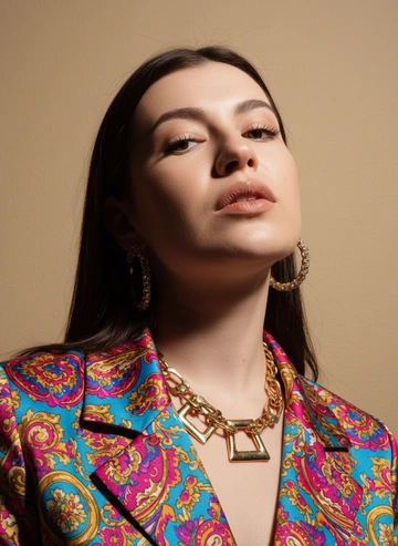 Создай портрет, используя те же черты лица (на основе загруженного фото), характеризующийся резким кинематографическим освещением и интенсивным контрастом. Снятый с немного нижнего ракурса, направленного вверх, что подчеркивает линию подбородка и шеи модели, этот портрет производит впечатление доминантности и скульптурной элегантности. Фон — бежевый. Яркая одежда. Добавить аксессуары. Портрет не в полный рост.