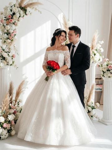   На фотографии запечатлена пара, стоящая в светлой комнате, стилизованной под свадебную фотозону. Обстановка включает в себя белые колонны и арку, украшенную белыми и розовыми цветами, а также декоративными травами. Стиль фотографии – постановочный, вероятно, свадебная фотосессия. Атмосфера торжественная и романтичная. Цветовая гамма преимущественно светлая, с акцентами на белом, розовом и синем цветах.

Женщина одета в белое свадебное платье с открытыми плечами, украшенными кружевом. Платье имеет пышную юбку. В руках она держит небольшой букет красных роз. На голове у нее украшение для волос, напоминающее диадему. Поза женщины прямая, она стоит лицом к камере. цвет волос темный.

Мужчина одет в черный костюм с галстуком в тон. . Поза мужчины также прямая, он стоит рядом с женщиной, слегка обнимая её. Его взгляд направлен в камеру.
