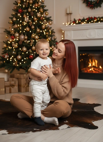 Молодая женщина европейской внешности и маленький ребенок, сидящие на полу, обнимаются в теплой рождественской обстановке. Женщина, приблизительно 28 лет, женщина одета в светло-коричневый свитер и брюки. Она обнимает маленького мальчика, около 2 лет, со светлыми волосами, который одет в белую футболку и белые. Оба улыбаются. Мальчик смотрит в камеру, а женщина в сторону ребенка. Они сидят, скрестив ноги, на полу из дерева, на ковре в форме шкуры коровы. Позади них — украшенная рождественская елка с гирляндами и огнями. Справа — камин с огнями. Свет исходит от огней на елке и камине, создавая теплую и уютную атмосферу. Композиция вертикальная, перспектива – средний план, подчеркивает близость и теплоту отношений между матерью и сыном. Преобладающие цвета – теплые оттенки коричневого, золотистого и зеленого. Не меняй черты лица и цвет волос
