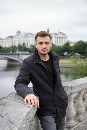 Мужчина в темном пиджаке стоит у гранитного парапета набережной, ладонь лежит на ограждении, он уверенно смотрит прямо в камеру. Позади — спокойная река, аксессуары городской архитектуры и зелень, рассеянный дневной свет, средний план.
