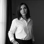 Женщина с блондинистыми волосами в чёрном топе и джинсах, позирует у серого здания.